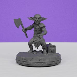 May include: A gray miniature figurine of a goblin holding an axe, with a sinister grin. The goblin is standing on a circular base with a treasure chest. The figurine is detailed and appears to be 3D-printed.