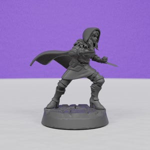 May include: A gray figurine of a female warrior in a hooded cloak, holding a sword. The figure is posed in a dynamic stance, with detailed armor and boots. The base is a textured, circular platform.