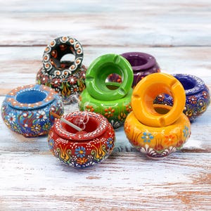 Può includere: Una collezione di colorati posacenere in ceramica con intricati motivi floreali. I posacenere sono in vari colori, tra cui rosso, blu, verde, giallo e viola. Un posacenere ha una sigaretta accesa. I posacenere sono rotondi con un'apertura circolare.