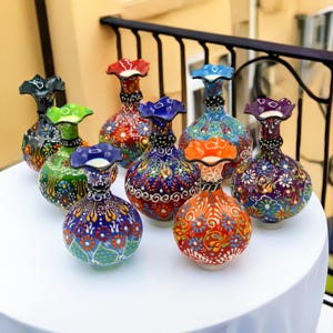 May include: Eight colorful ceramic vases with intricate floral patterns. The vases are all different colors and designs, and they are arranged in a circle on a white surface.