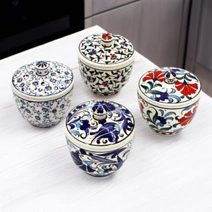 Puede incluir: Cuatro tarros de cerámica con tapa y diseños florales. Los tarros son blancos con diseños azules, rojos y verdes. Cada tarro tiene un pequeño pomo en la tapa. Los tarros están dispuestos sobre una superficie blanca.