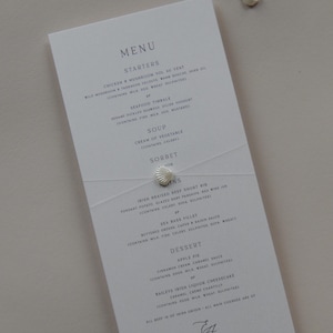 May include: A white menu card listing starters, soup, sorbet, and mains. The menu is tied with a white ribbon and a seashell. The text is in a simple, elegant font. The menu is for a wedding or special event.