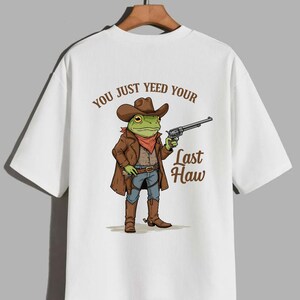 May include: White t-shirt with a cartoon frog wearing a cowboy hat and holding a gun. The text "You just yeed your last haw" is printed on the back of the shirt.