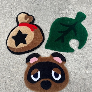 May include: Three plush rugs featuring characters from the Animal Crossing video game series. The rugs are brown, green, and tan. The brown rug features the face of Tom Nook, the green rug features a green leaf with a nook on it, and the tan rug features a money bag with a star on it.