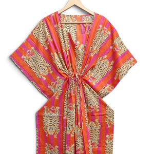Op de afbeelding: Een levendige kaftan met een herhalend tijgerpatroon tegen verticale strepen van feloranje en felroze. Het kledingstuk heeft een V-hals, wijde mouwen in kimonostijl en een trekkoord in de taille, getoond op een houten kledinghanger.