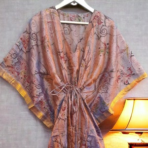 May include: A long, flowing, pink and purple patterned kaftan with gold trim. The kaftan is hanging on a hanger in front of a light-coloured wall.