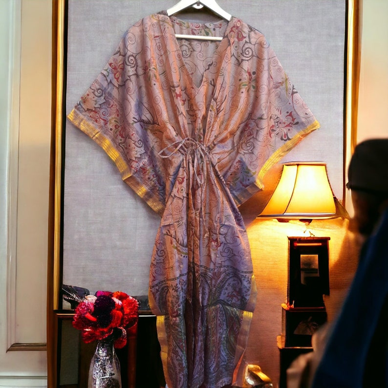 May include: A long, flowing, pink and purple patterned kaftan with gold trim. The kaftan is hanging on a hanger in front of a light-coloured wall.