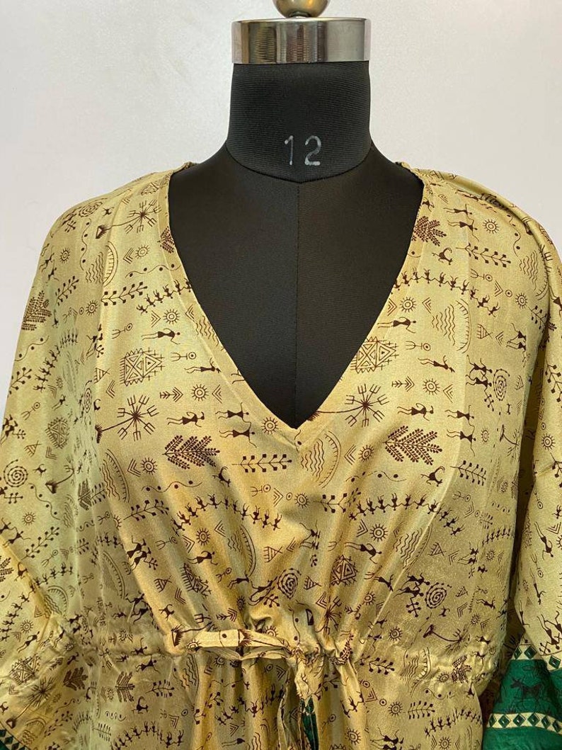 Op de afbeelding: Een beige kaftan met een V-hals en een strik in de taille. De kaftan is bedekt met een bruin en groen patroon van abstracte vormen en figuren.