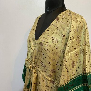 Op de afbeelding: Een beige zijden kaftan met een groene bies en een bruin abstract patroon. De kaftan heeft een V-hals en een strik in de taille.