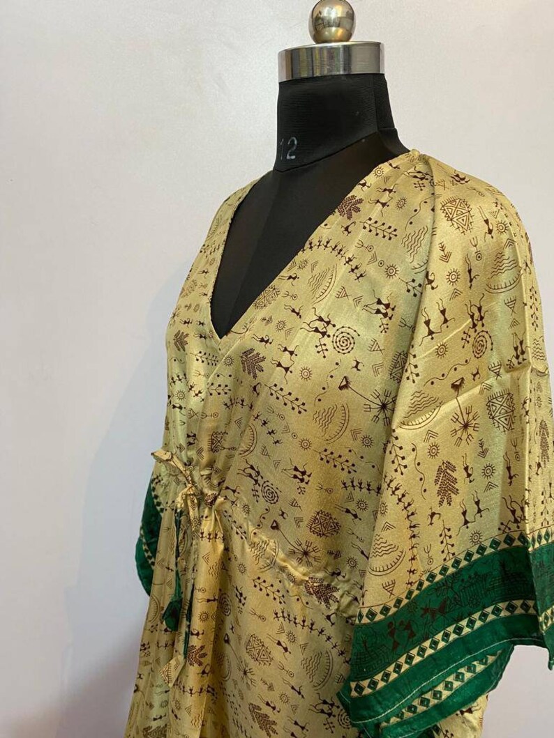 Op de afbeelding: Een beige zijden kaftan met een groene bies en een bruin abstract patroon. De kaftan heeft een V-hals en een strik in de taille.