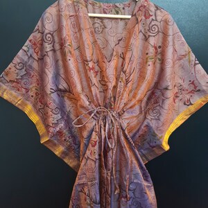 May include: A pink and brown patterned kaftan with gold trim. The kaftan has a V-neck and a drawstring waist.