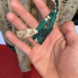 Op de afbeelding: Close-up van een groene en beige stoffen stropdas met een geometrisch patroon. De stropdas is in een knoop geknoopt en wordt vastgehouden door een hand.