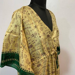 Op de afbeelding: Een lange, vloeiende, gele zijden kaftan met een groene bies en een bruine tribale print. De kaftan heeft een V-hals en een strik in de taille.