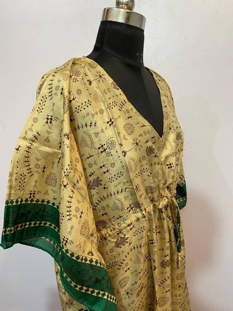 Op de afbeelding: Een lange, vloeiende, gele zijden kaftan met een groene bies en een bruine tribale print. De kaftan heeft een V-hals en een strik in de taille.