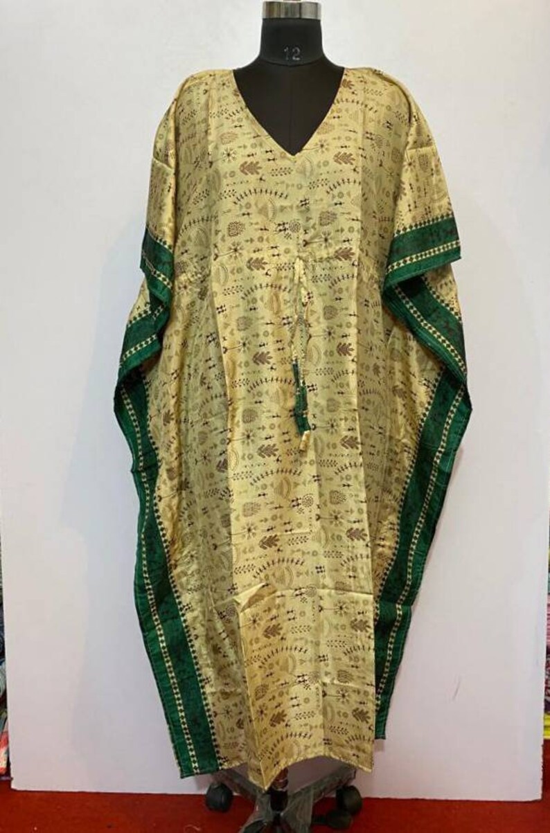 Op de afbeelding: Een lange, beige kaftan met een groene bies en een geometrisch patroon. De kaftan heeft een V-hals en een trekkoord in de taille.
