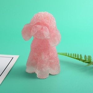 Op de afbeelding: Een roze poedelvormige kaars op een lichtgroene achtergrond. De kaars is gemaakt van een doorschijnend materiaal en heeft een glinsterende afwerking.