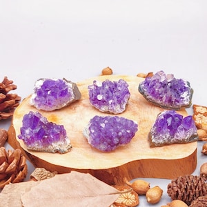 May include: Six amethyst crystal clusters of varying sizes are arranged on a wooden slab. The crystals display a deep purple hue. The arrangement is surrounded by pine cones, dried leaves, and other natural elements, creating a rustic aesthetic.