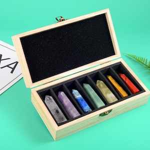 Może przedstawiać: Drewniana skrzynka z zawiasowym wiekiem, zawierająca siedem kryształowych różdżek w różnych kolorach. Różdżki są ułożone w rzędzie, z kolorami od fioletowego do czerwonego. Skrzynka jest wyłożona czarnym aksamitem.