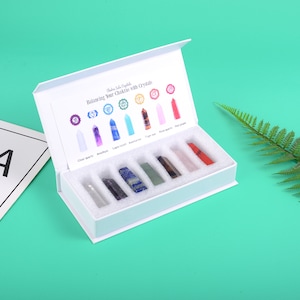 Op de afbeelding: Een witte doos met een open deksel, waarin een set van zeven kristallen punten in een witte schuimrubberen inlegger te zien is. De doos heeft een label met de tekst "Chakra Jade Crystals Balancing Your Chakras with Crystals".