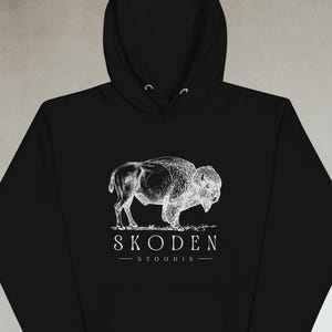 May include: Black hoodie featuring a white illustration of a bison. The image includes the text "SKODEN - STOODIS-" below the bison. The hoodie has a drawstring hood and a front pocket.