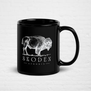 May include: Black ceramic mug featuring a white illustration of a bison. The mug has the text "SKODEN - STOODIS -" in white. The design is a detailed, vintage-style drawing of the animal.