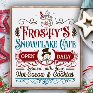 以下が含まれることがあります： 冬のテーマの素朴な木製の看板。看板には「フロスティのスノーフレイクカフェ」と雪だるまのイラスト、そして「毎日営業」と「愛情込めて提供 - ホットココア＆クッキー」の文字が書かれています。