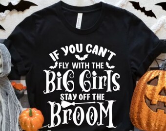 Può includere: T-shirt nera con la scritta bianca "If You Can't Fly With The Big Girls Stay Off The Broom". L'immagine include decorazioni di Halloween, come pipistrelli, zucche e un teschio.