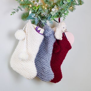 May include: Three knitted Christmas stockings hanging from a green garland. The stockings are white, gray, and red. The white stocking has a white pom-pom on the top and a small white gift inside. The gray stocking has a white pom-pom on the top. The red stocking has a white pom-pom on the top and a small stuffed animal inside.