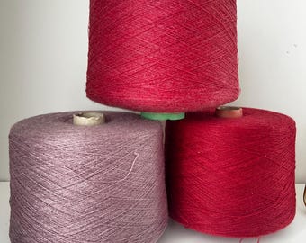 May include: Three spools of yarn in shades of red and purple. The top spool is a vibrant red, the bottom right is a darker red, and the bottom left is a muted purple. The yarn is tightly wound on cardboard cores, ready for crafting projects.