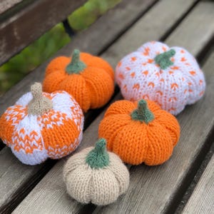 May include: Five knitted pumpkins in various sizes and colours. The pumpkins are orange, white, and beige. They are sitting on a wooden surface.