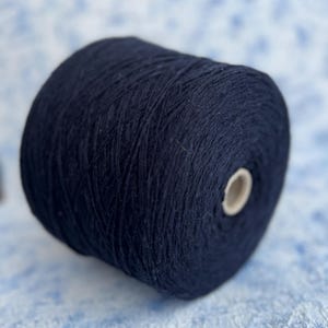 Peut inclure: Une grosse bobine de fil bleu marine. Le fil est étroitement enroulé, montrant une surface texturée. Le cœur de la bobine est visible à une extrémité. L'arrière-plan est un motif moucheté bleu clair et blanc.