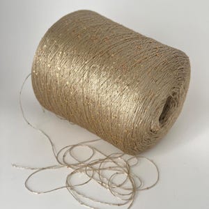 May include: A spool of tan-colored yarn with gold sequins. The yarn is tightly wound, and a strand of the yarn extends from the spool. The yarn is suitable for knitting, crocheting, or other craft projects.
