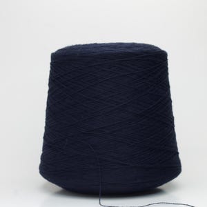 May include: A large cone of navy blue yarn, tightly wound and ready for crafting. The yarn is a deep, solid colour, and a strand extends from the cone. The yarn is set against a plain white background.
