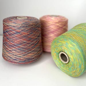 Peut inclure: Trois cônes de fil colorés. Le premier est un mélange de fils rouges, bleus et beiges. Le deuxième est rose et orange. Le troisième est vert et jaune. Le fil est enroulé autour de tubes en carton.