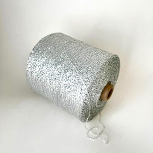 May include: A silver metallic yarn spool. The yarn is tightly wound around a brown cardboard core. The yarn has a shiny, textured appearance, and a strand of yarn extends from the spool.