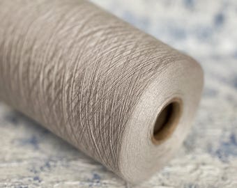 Puede incluir: Una bobina de hilo de color gris claro con un brillo sutil. El hilo está enrollado apretado y tiene una textura suave.