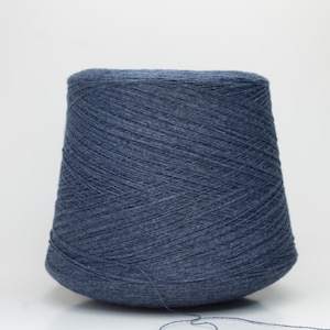May include: A large cone of denim blue yarn, tightly wound, is positioned on a white surface. The yarn has a solid colour with a slightly textured appearance. The cone tapers from a wide base to a rounded top. A strand of yarn extends from the cone.