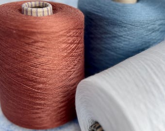 May include: Close-up of three spools of yarn in shades of rust, blue, and white. The yarn is tightly wound around cardboard cores. The rust-colored spool is in the foreground, with the blue and white spools behind it. The yarn has a slight sheen.