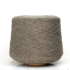 May include: A large cone of grey yarn, tightly wound and ready for crafting. The yarn is a neutral shade, suitable for various projects. The cone sits on a wooden base, with a strand extending from the top.