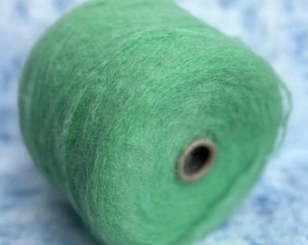 May include: A close-up of a cone of green yarn. The yarn has a soft, fuzzy texture and is wound around a cardboard core. The yarn is a light, spring green colour, and the background is a light blue.
