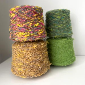 May include: Four cones of colourful yarn are stacked. The top cone is a mix of pink, yellow, and grey. Below it is a brown cone with yellow flecks. The other two cones are green, ideal for knitting or crochet.
