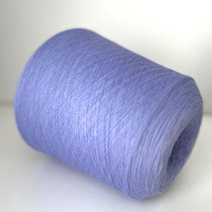 Peut inclure: Une grosse bobine de fil de couleur lavande. Le fil est étroitement enroulé, créant une forme cylindrique. La texture du fil est visible, avec des brins individuels créant une surface légèrement texturée. La bobine est posée sur un fond blanc uni.