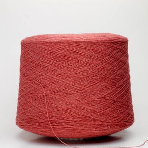May include: A large spool of coral-coloured yarn. The yarn is tightly wound, creating a textured surface. A single strand of yarn extends from the spool. Ideal for knitting, crocheting, or other fibre arts projects.