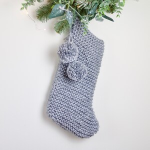 May include: A gray knitted stocking with two fluffy pom-poms at the top. The stocking is hanging from a green garland.