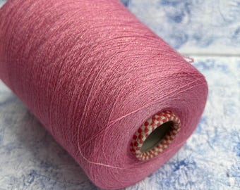 May include: A large spool of pink yarn. The yarn is tightly wound and has a slight sheen. The core of the spool is visible, featuring a red and white checkered pattern. The background is a textured, light blue and white surface.