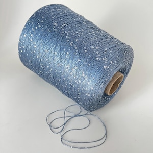 May include: A large spool of blue yarn with silver sequins. The yarn is wound around a cardboard core, with a length of the yarn unwound and lying on the surface. The yarn has a slight sheen and is suitable for crafting projects.