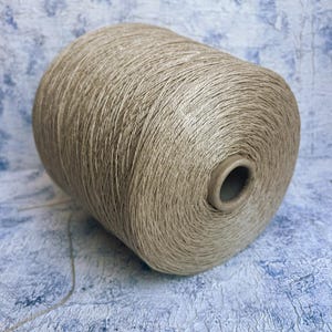 May include: A spool of beige yarn, likely linen, with a natural, slightly rough texture. The yarn is wound tightly on the spool, which is made of cardboard.
