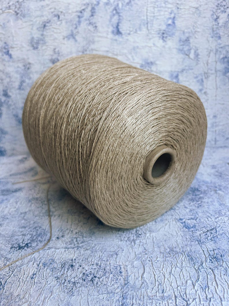 May include: A spool of beige yarn, likely linen, with a natural, slightly rough texture. The yarn is wound tightly on the spool, which is made of cardboard.