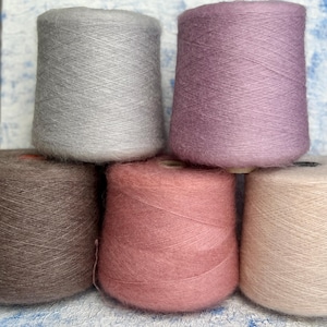 May include: A collection of six yarn cones in various soft colors. The colors include gray, lavender, brown, dusty rose, and cream. The yarn appears to be a soft, fluffy material, ideal for knitting or crocheting projects.