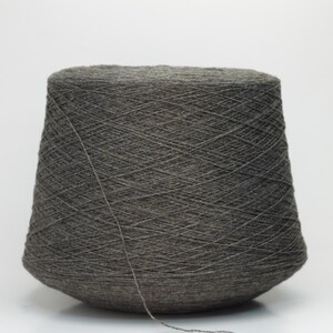 May include: A large cone of dark grey yarn, ideal for knitting or crochet projects. The yarn is tightly wound, with a visible texture and a single strand extending from the cone. The background is plain white.